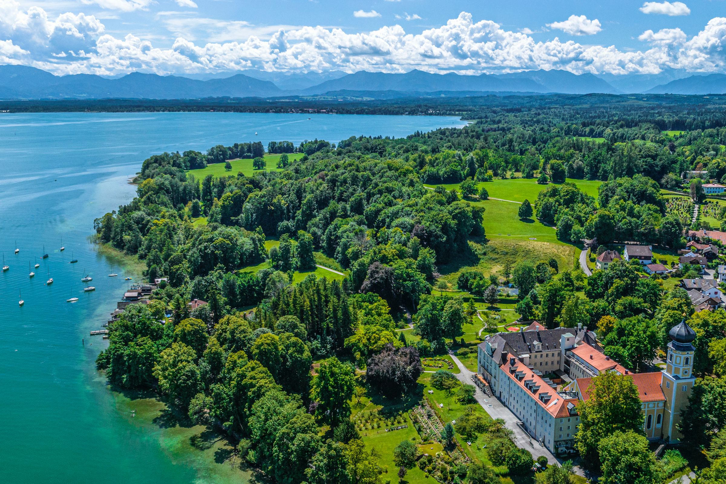 Luftbild des Bernrieder Parks: Links liegt der intensiv blaue Starnberger See, rechts der grüne Park. Im Hintergund sind die Alpen zu sehen.
