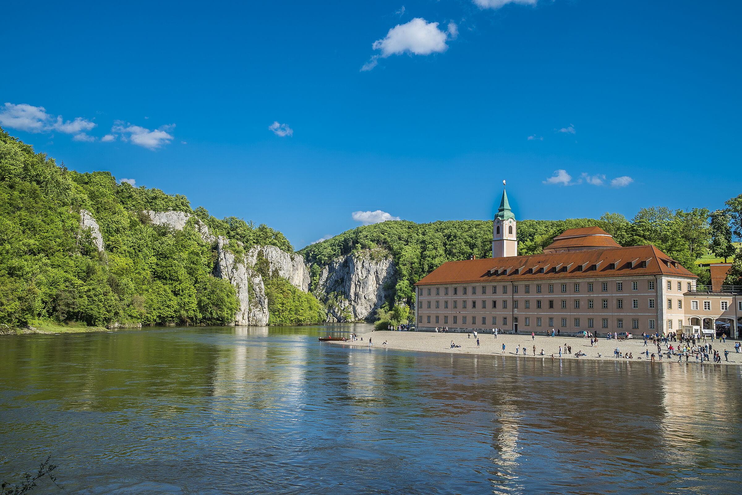 Das Kloster Weltenburg an einem sonnigen Tag, das Gebäude spiegelt sich in der Donau, die Hänge des Donaudurchbruchs sind bewaldet, der Wald wird von den charakteristischen Felsen durchbrochen