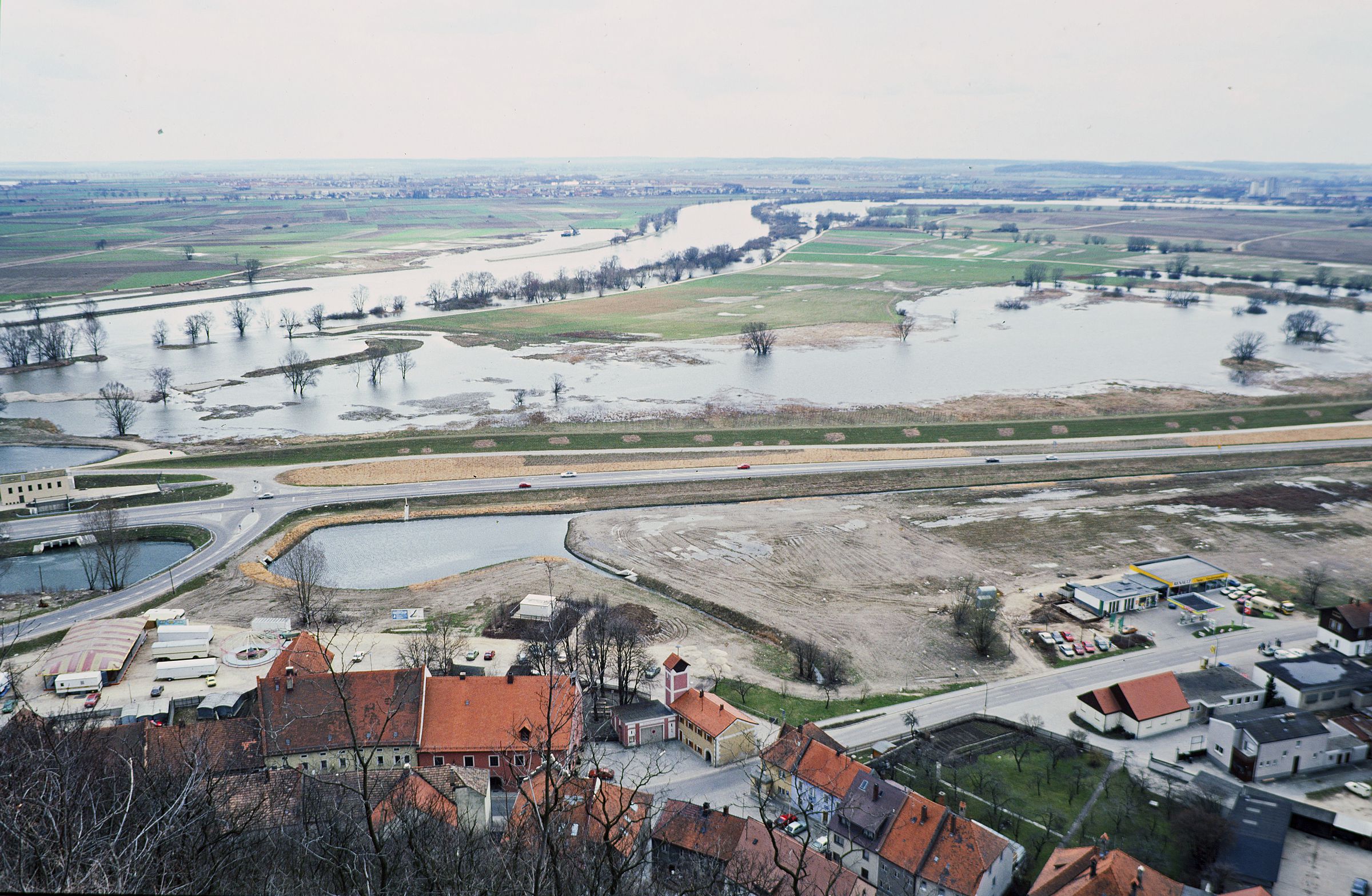 Ein Bild auf das Donaustaufer Altwasser im Jahr 1986. Eine Schnellstraße zerteilt den Fluss und das Altwasser. Neben der Straße ist eine Wohnsiedlung, planiertes Bauland und einige Parkplätze. Auf der anderen Seite der Donau sind Felder und der nächste Ort zu erkennen 