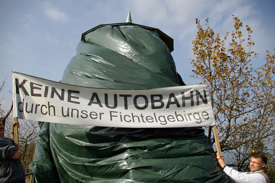 Eine dunkelgrüne Plane verhüllt den Aussichtsturm auf dem Waldstein. Davor stehen zwei Menschen mit einem Plakat, auf dem "KEINE AUTOBAHN durch unser Fichtelgebirge" zu lesen ist.