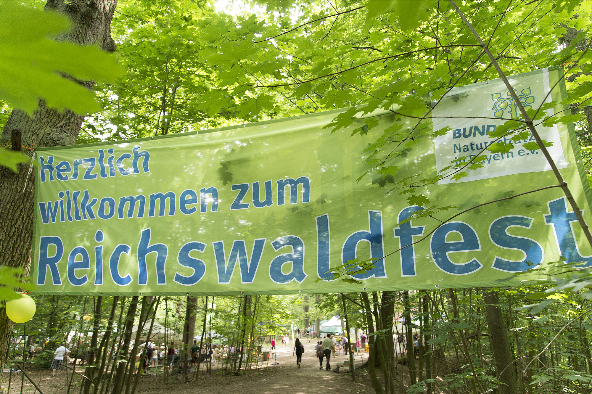 Auf einem großen grünen, zwischen Bäumen aufgespannten Banner steht "Herzlich willkommen zum Reichswaldfest"