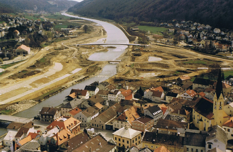 Eine graue und braune, unnatürliche Landschaft. Der Fluss ist begradigt und die Ufer sind aufgegraben. Wo keine Baustelle ist, da stehen Häuser. 