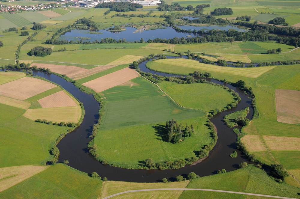 Eine Luftaufnahme der Regentalaue und des Rötelseeweihers: ein zwischen kleinen Feldern mäandernder Fluss und ein eckiger Weiher