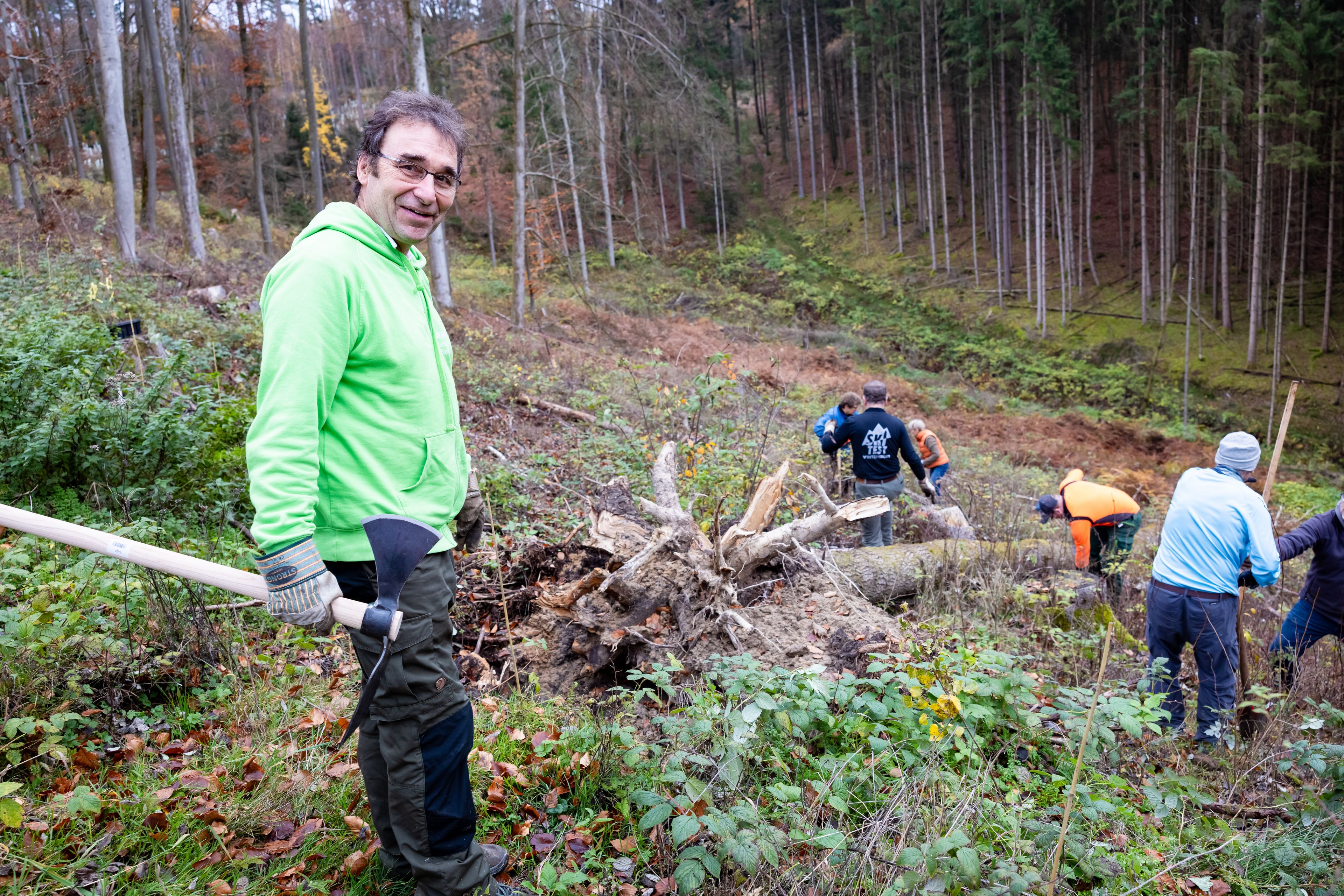 Foto: Jörg Farys Der BN Experte Ralf Straßberger im Vordergrund mit einer Hacke, im Hintergrund Ehrenamtliche auf einer unbewaldeten Fläche. Es werden Bäume gepflanzt