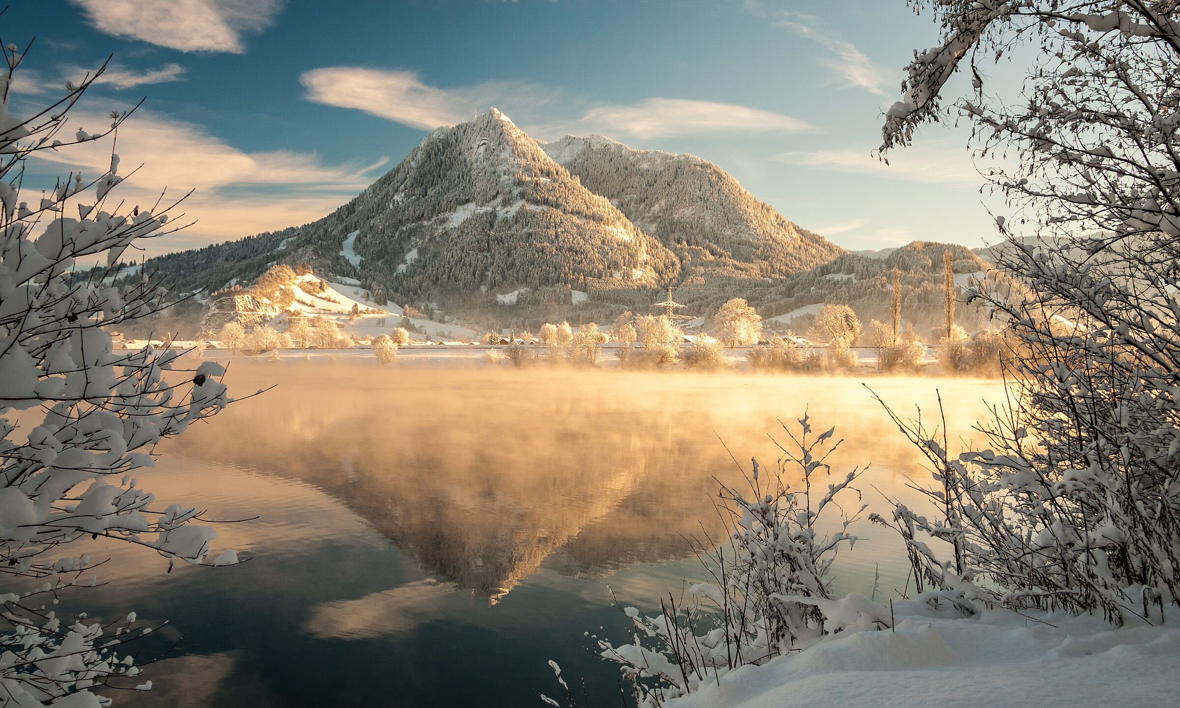 Der schneebedeckte Grünten im Winter erstrahlt in schrägem Sonnenlicht. Im Vordergrund liegt ein See, aus dem Nebel aufsteigt.