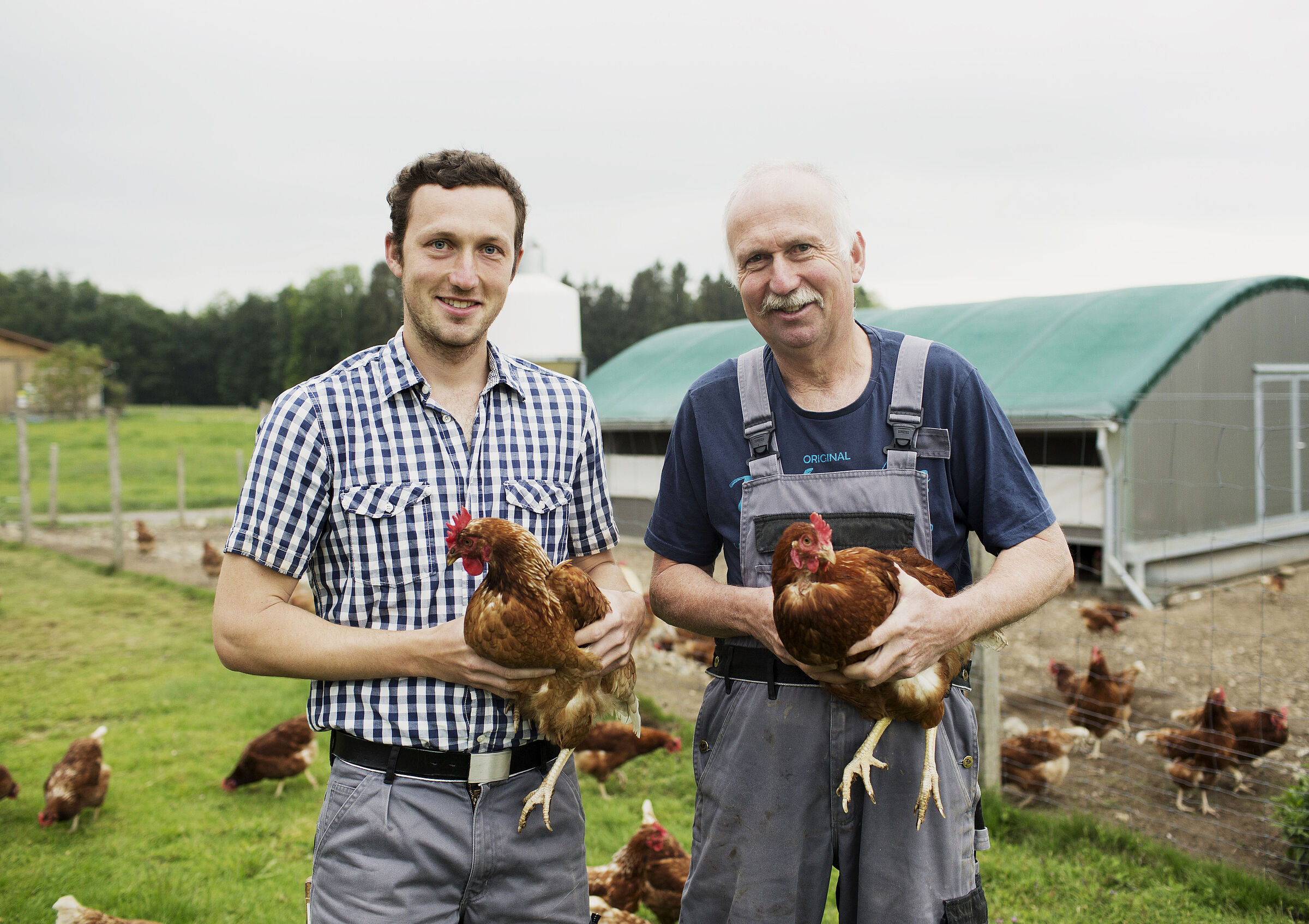 Junior- und Seniorbauer in Arbeitskleidung mit je einem Huhn auf dem Arm vor ihrem Hühner-Freilauf