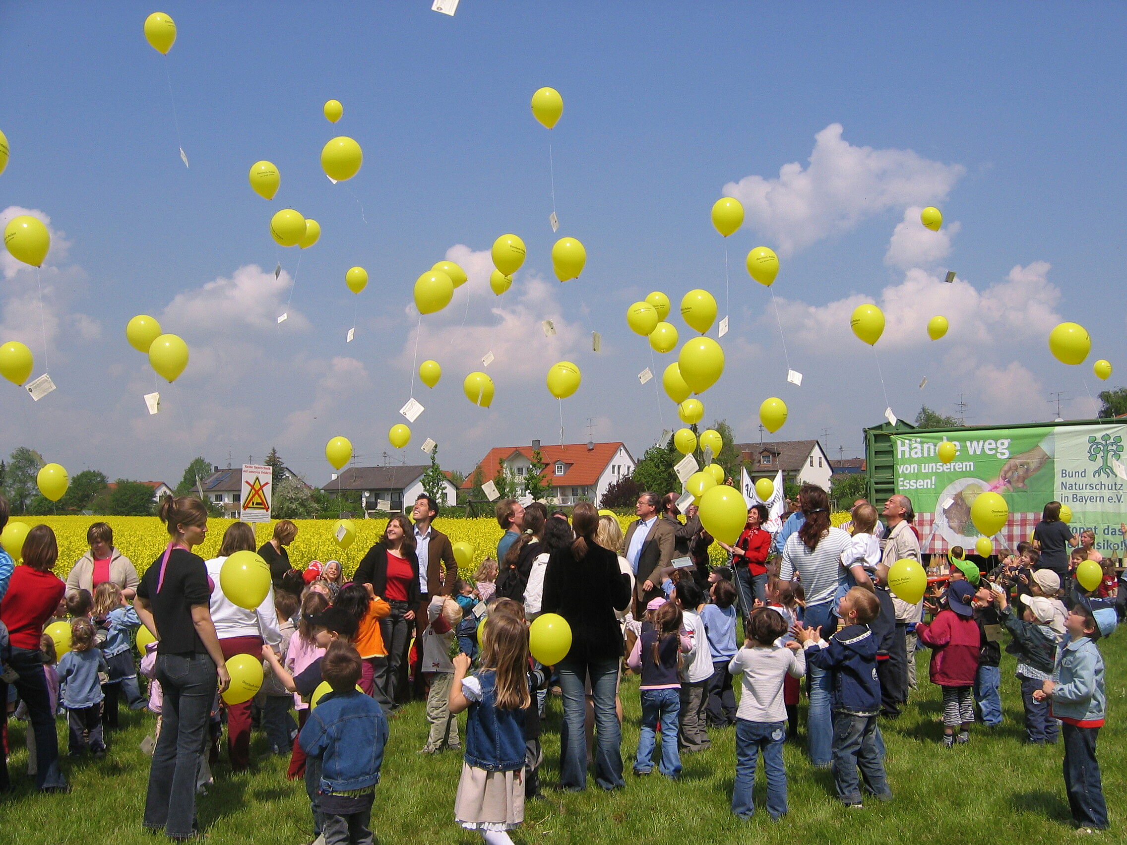 Demonstration gegen Gentechnik in der Landwirtschaft: Erwachsene und viele Kinder stehen vor einem Rapsfeld und lassen gelbe Luftballons in den Himmel steigen.