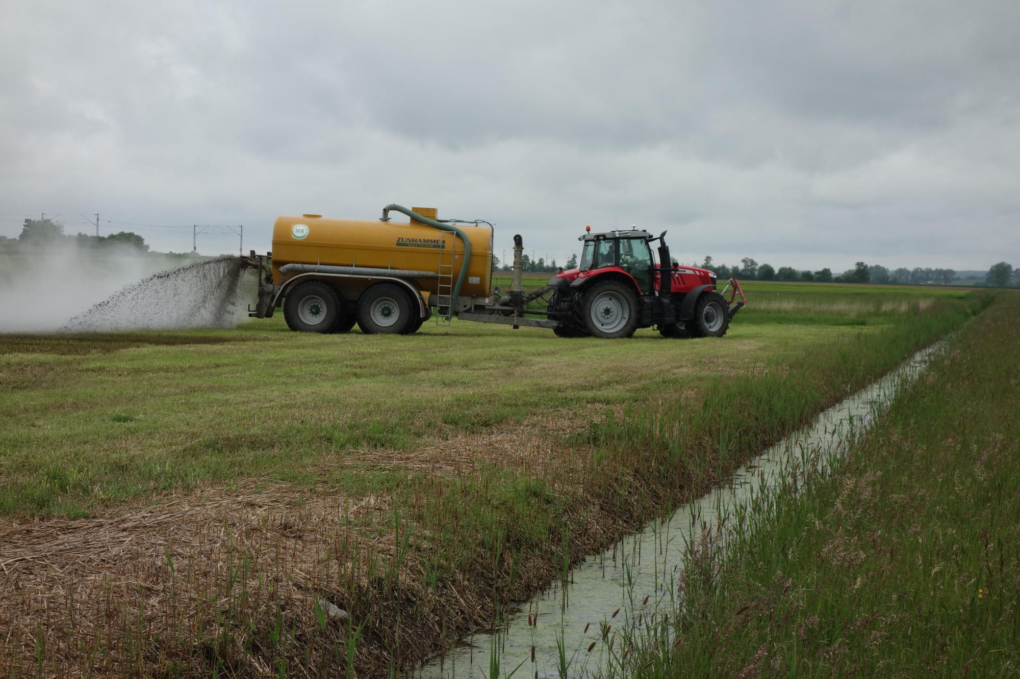 Nitrat im Grundwasser: Ein Traktor verteilt auf einer Wiese Guelle neben einem Wasserlauf