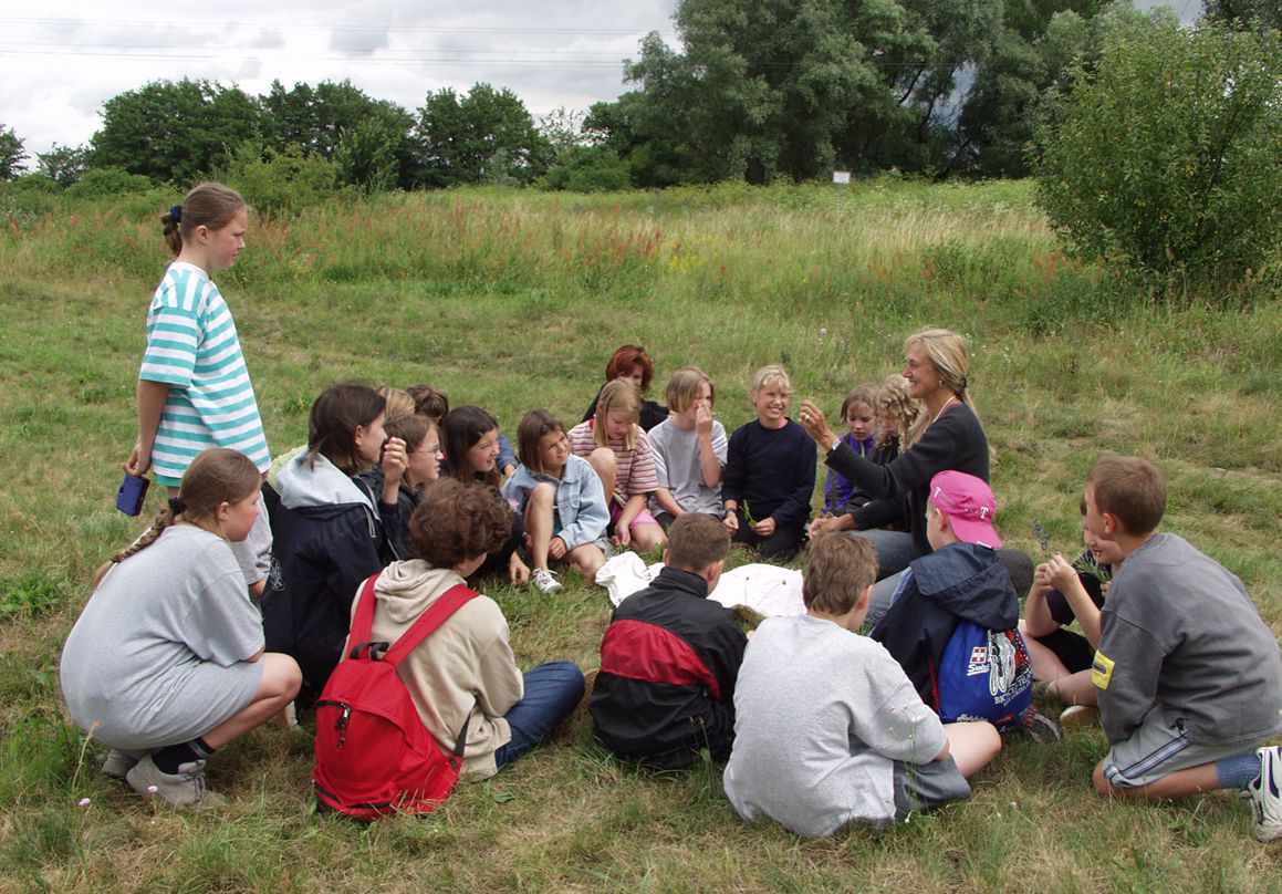 Umweltbildungs-Veranstaltung zum Thema Sand – eine Gruppe Kinder sitzt mit einer Pädagogin im Kreis auf einer Wiese.