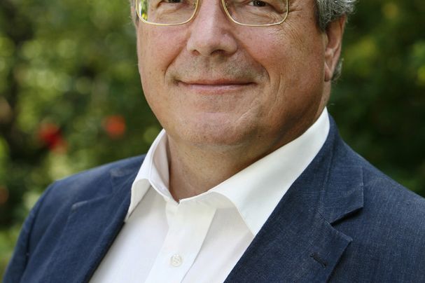 Das Portrait eines Mannes mit weißen Haaren und Brille in einem weißen Hemd und einer blauen Anzugjacke.