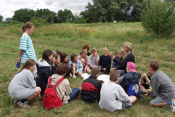 Umweltbildungs-Veranstaltung zum Thema Sand – eine Gruppe Kinder sitzt mit einer Pädagogin im Kreis auf einer Wiese.