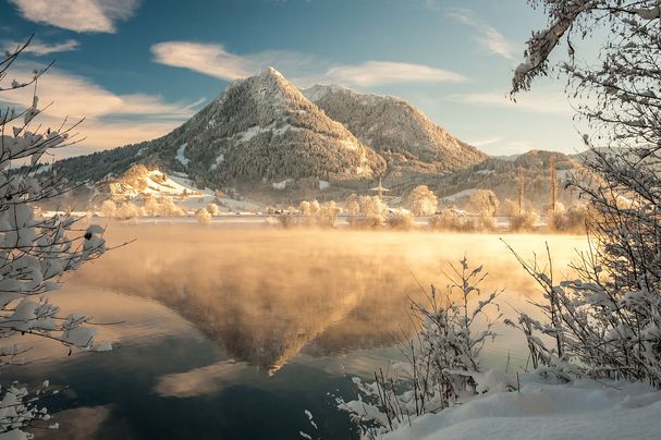 Der schneebedeckte Grünten im Winter erstrahlt in schrägem Sonnenlicht. Im Vordergrund liegt ein See, aus dem Nebel aufsteigt.