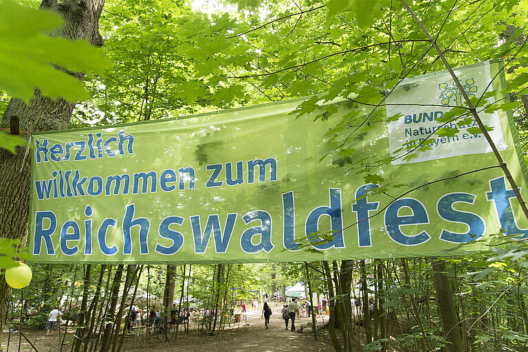 Im Wald hängt ein großes grünes Banner mit der Aufschrift "Herzlich willkommen zum Reichswaldfest"