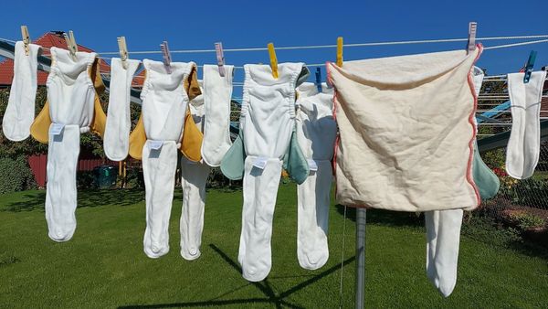 Mit der richtigen Wahl und Anwendung von Stoffwindeln fällt deren Umweltbilanz deutlich besser aus als die von Einwegwindeln. (Foto: Susann Koethe/stock.adobe.com) Stoffwindeln richtig nutzen: Einige weiße Stoffwindeln hängen an einer Wäscheleine, um zu trocknen. Im Hintergrund sieht man einen grünen Garten und blauen Himmel.
