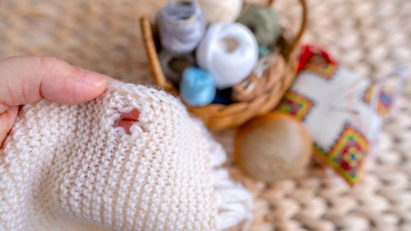Kleidung möglichst lange zu verwenden und bei Bedarf zu reparieren, trägt viel zur Nachhaltigkeit bei. (Foto: kittyfly/stock.adobe.com) Nachhaltige Kleidung: Eine Hand hält einen hellen Wollpulli mit einem Loch. Im Hintergrund sieht man Material, um das Loch zu flicken.