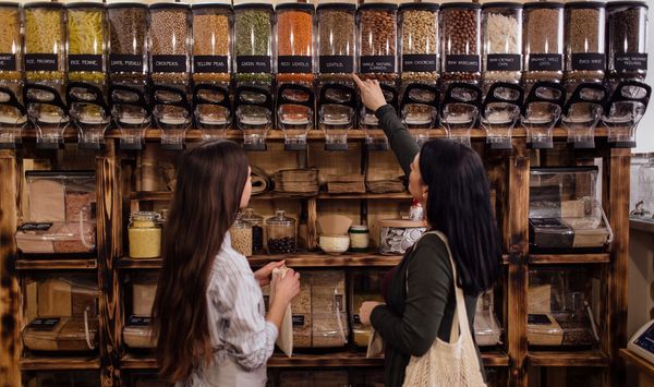 Umweltfreundliche Verpackungen: Am besten kauft man unverpackt bzw. nimmt wiederverwertbare, eigene Behälter mit. (Foto: Newman Studio/stock.adobe.com) Umweltfreundliche Verpackungen: zwei Frauen stehen in einem Zero-Waste-Laden vor großen, durchsichtigen Behältern mit verschiedenen Lebensmitteln.