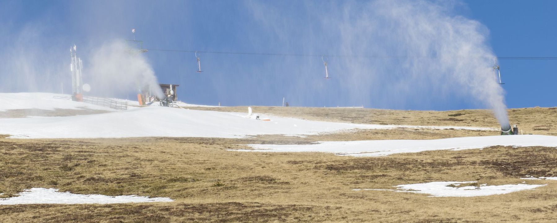 Die Kosten des Alpentourismus: Schneekanonen in Betrieb an einem nahezu schneefreien Hang im Sonnenlicht. (Foto: AdobeStock/mg1708) 