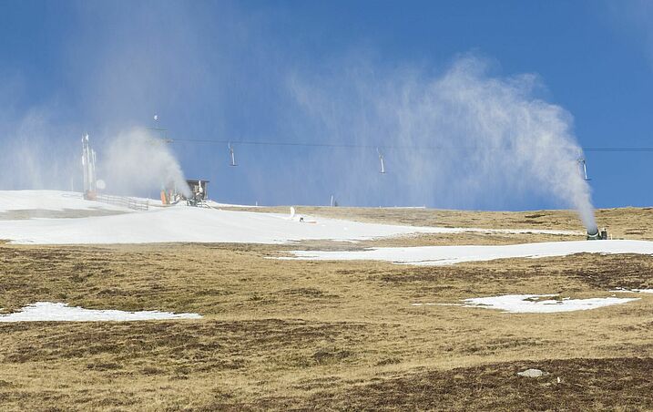Immer mehr Flächen werden für den Alpinskisport künstlich beschneit, daneben findet sich immer öfter grüne Landschaft. (Foto: AdobeStock/mg1708) Schneekanonen in Betrieb an einem nahezu schneefreien Hang im Sonnenlicht.
