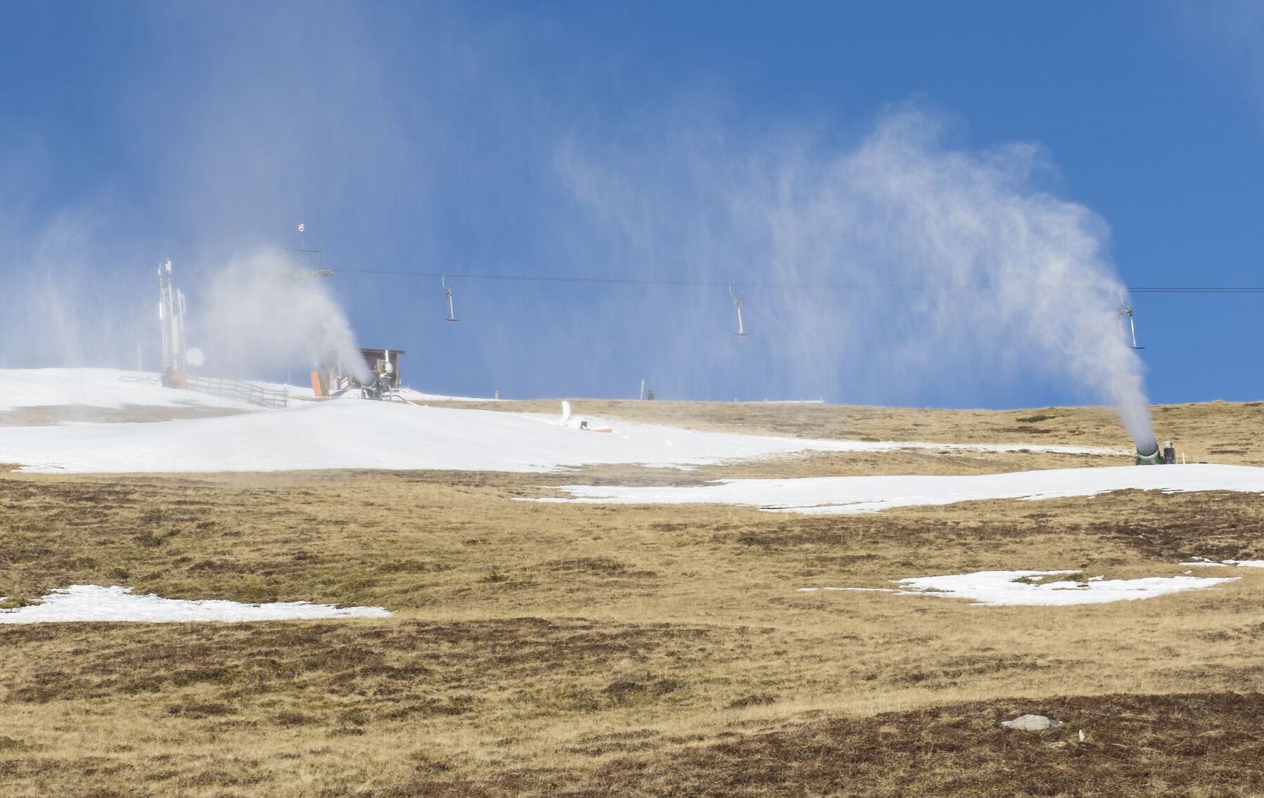 Schneekanonen in Betrieb an einem nahezu schneefreien Hang im Sonnenlicht.