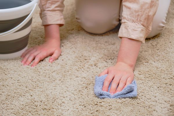 A woman with a bucket and a rag washes the carpet in the home living room Eine Frau kniet mit einem Lappen in der Hand auf einem hellen Boden, neben ihr ein Eimer. Leichte Flecken entfernt man mit Wasser oder Waschmittel.