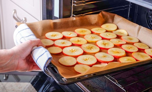 Lebensmittel konservieren: Beim Trocknen wird den Lebensmitteln Wasser entzogen, wodurch Keime keine Wachstumsgrundlage mehr haben. (Foto: Olga Gubskaya/stock.adobe.com) Lebensmittel konservieren: Apfelringe auf einem mit Papier abgedeckten Backblech, das gerade in den Ofen geschoben wird.