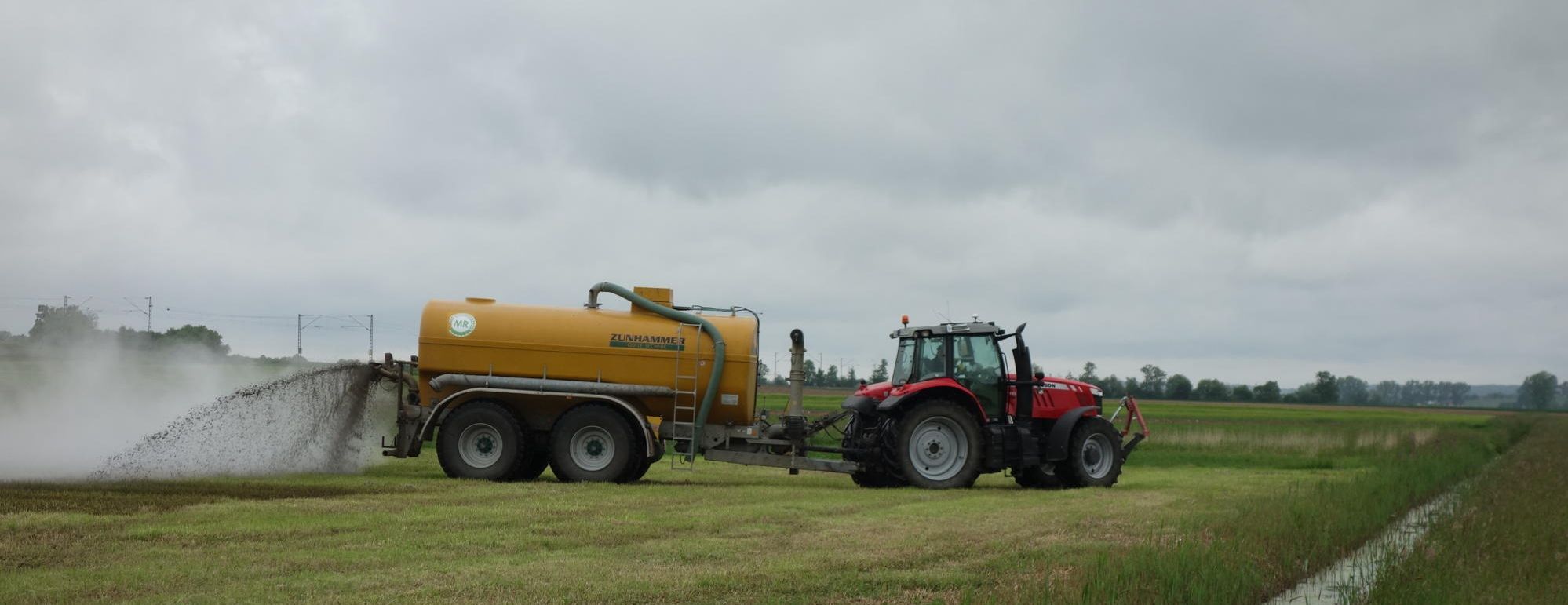 Nitrat im Grundwasser: Ein Traktor verteilt auf einer Wiese Guelle neben einem Wasserlauf
