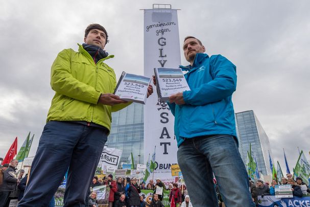 Zwei Männer halten Unterschriftslisten gegen Glyphosat in die Kamera, im Hintergrund sieht man Hochhäuser und Demonstranten mit Fahnen und Bannern (Foto: Toni Mader)