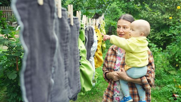 Stoffwindeln verursachen weniger Abfall, sind besser für die Haut und schonen den Geldbeutel. (Foto: Gargonia/stock.adobe.com) Stoffwindeln richtig nutzen: Eine Mutter mit ihrem Baby im Arm, steht vor einer Wäscheleine mit trocknenden Stoffwindeln. Im Hintergrund sieht man einen grünen Garten.