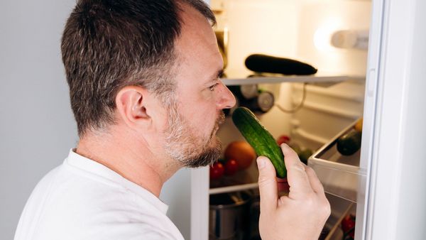 Lebensmittelverschwendung vermeiden: Ein Mann steht avor einem geöffneten Kühlschrank und riecht prüfend an einer Gurke.