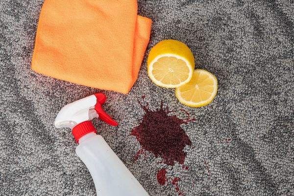 Foto: TSViPhoto/adobe.stock.de Ein roter Fleck auf einem grauen Teppich, daneben eine Sprühflasche, ein organger Lappen und zwei Zitronenhälften. Stärkere Flecken entfernt man mit Essig oder Zitronensaft.