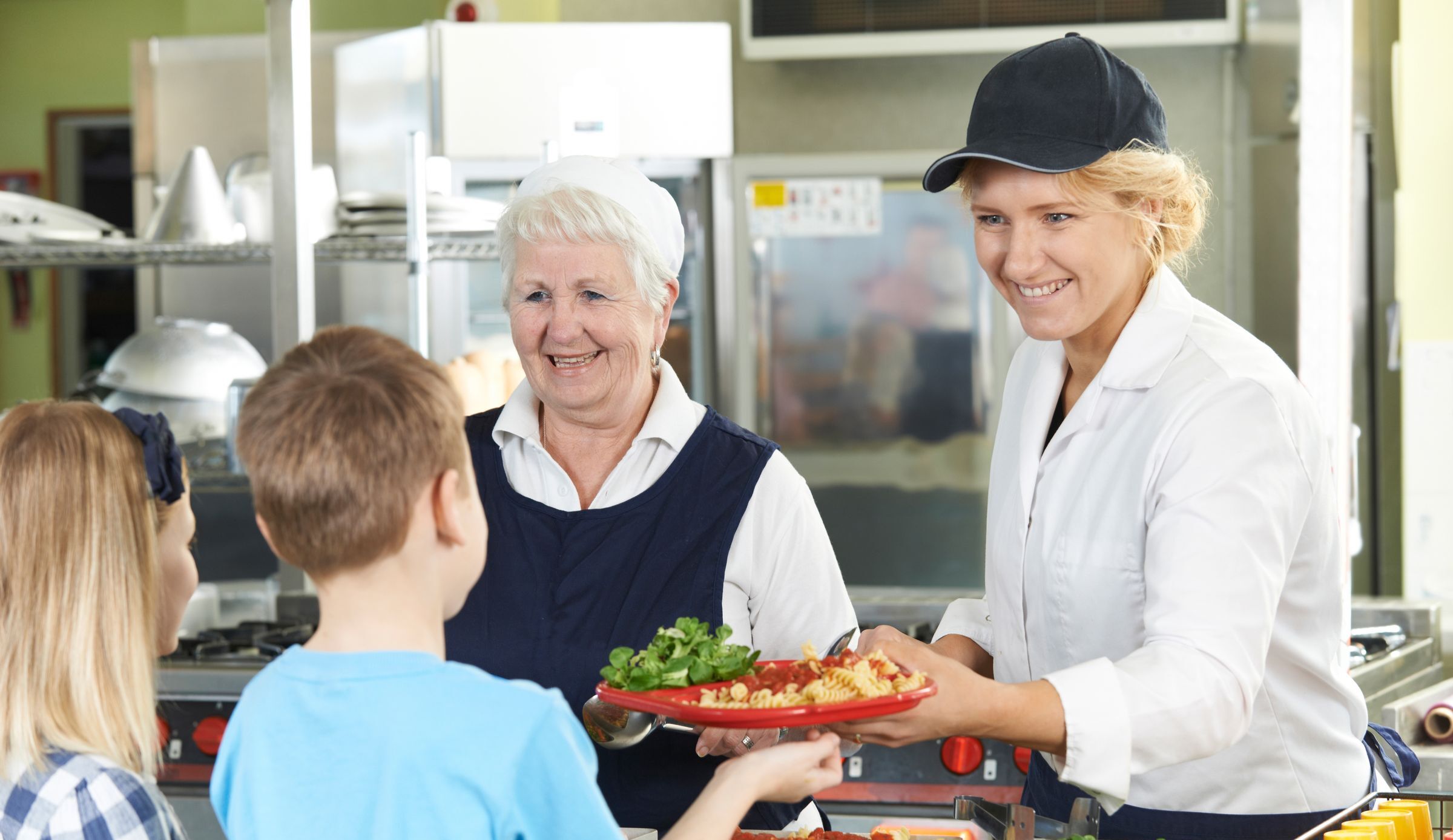 Biolandwirtschaft in Bayern: Zwei lächelnde Frauen teilen in einer Kantine Teller mit Nudeln und Salat an Kinder aus