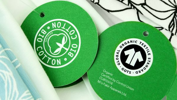 Für Kleidung gibt es etliche Siegel mit unterschiedlicher Aussagekraft. Das GOTS-Siegel bietet den umfassendsten Standard für Bio-Textilien und faire Sozialstandards. (Foto: Pixelot/stock.adobe.com) Nachhaltige Kleidung: Zwei grüne runde Anhänger mit dem GOTS-Siegel liegen auf einer Textilie.