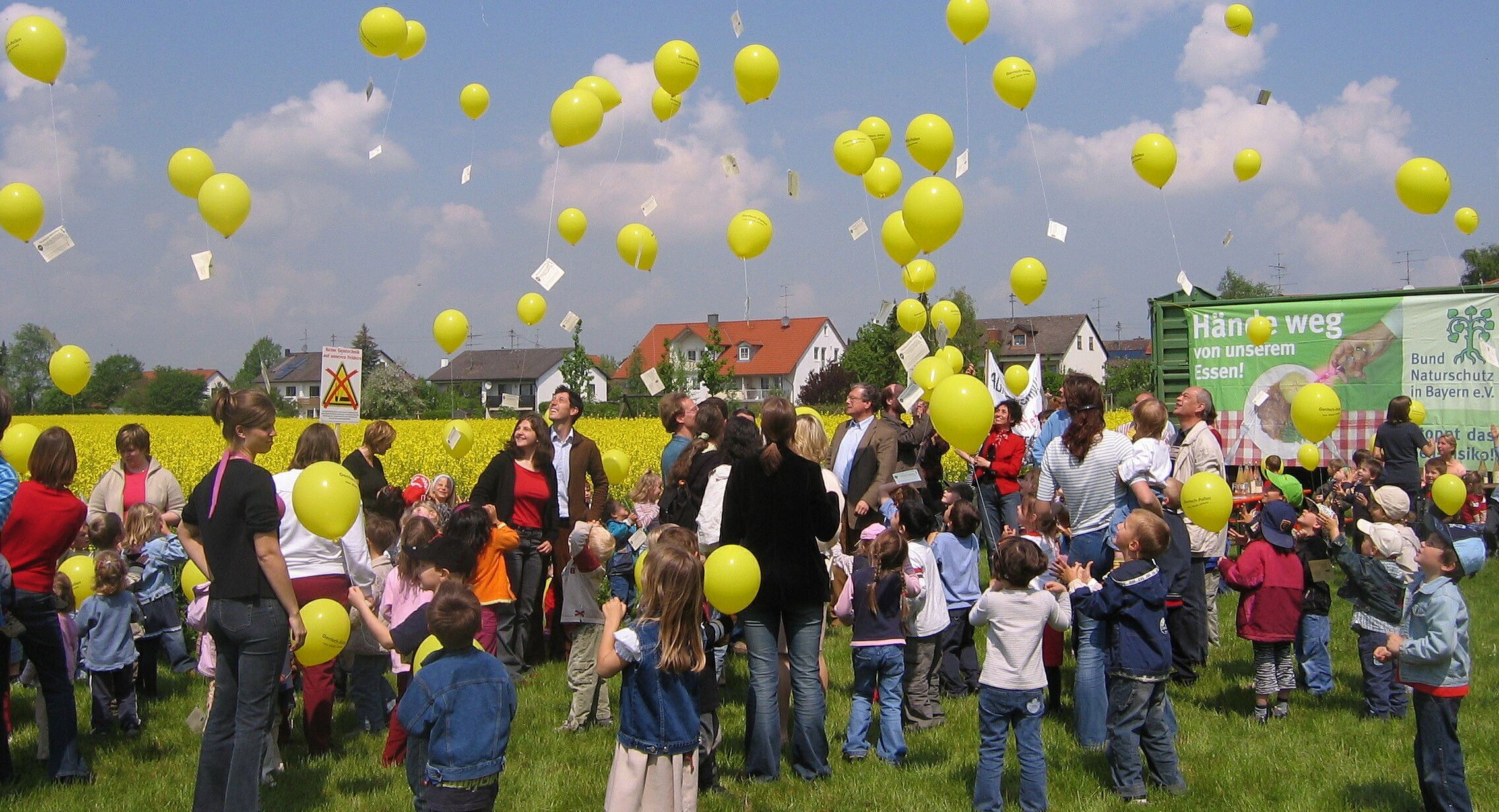 Luftballonaktion gegen Gentechnik in der Landwirtschaft. (Foto: Demonstration gegen Gentechnik in der Landwirtschaft: Erwachsene und viele Kinder stehen vor einem Rapsfeld und lassen gelbe Luftballons in den Himmel steigen.
