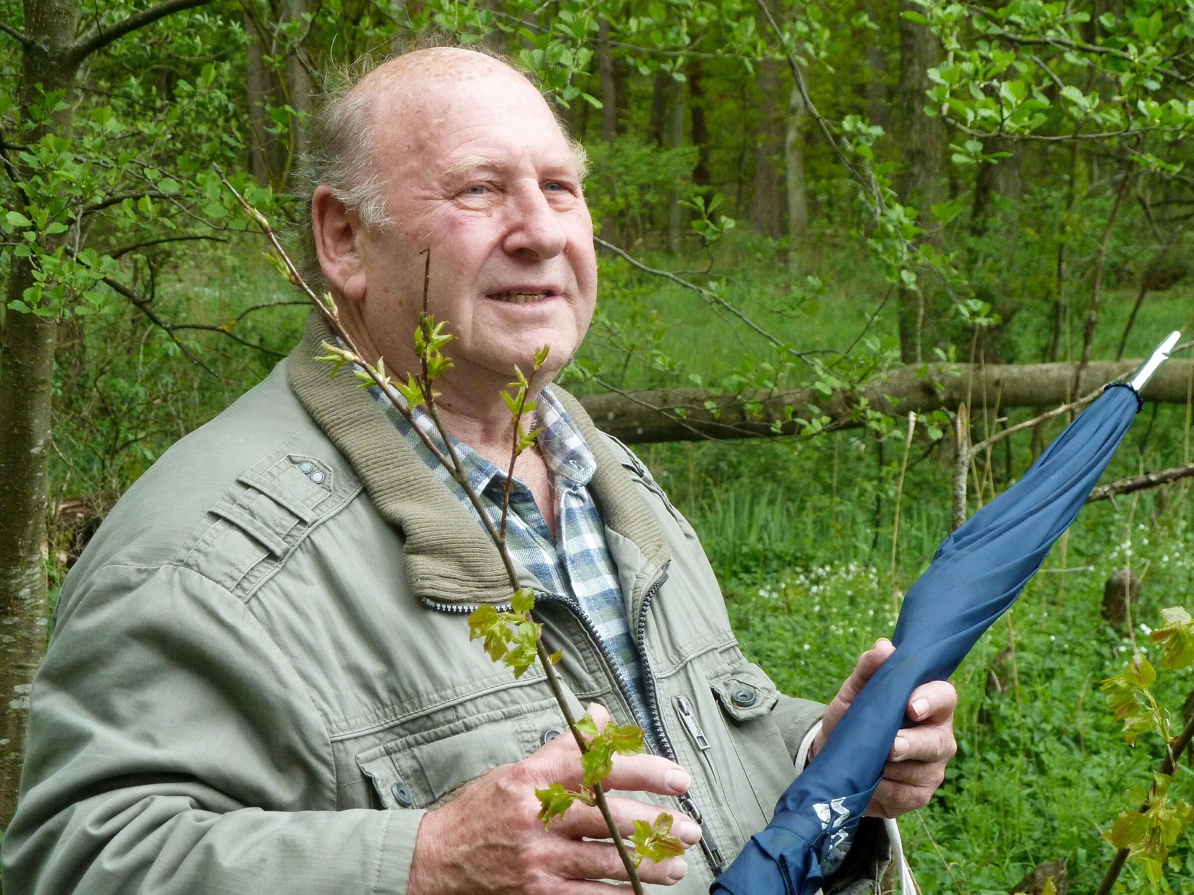Ein älterer Herr mit Stirnglatze, beiger Jacke, kariertem Hemd und blauem Regenschirm steht im Wald