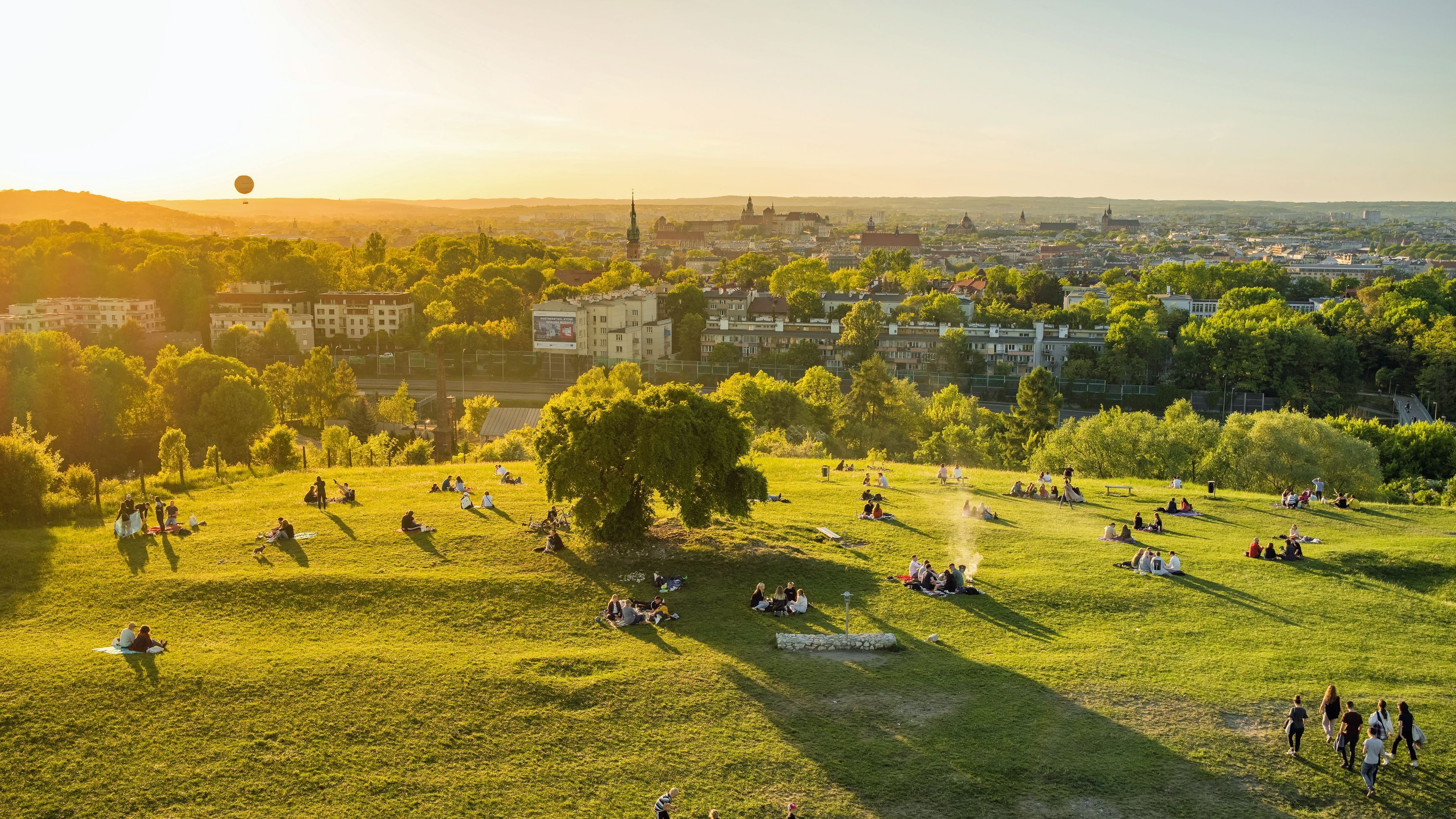 Stadtnatur ist eine wichtige Grundlage für die Gesundheit: Sie senkt das Stresslevel, steigert das Wohlbefinden, reinigt die Luft, kühlt überhitzte Straßen und fördert die Bewegung im Alltag (Foto: Mazur Travel/stock.adobe.com). Stadtnatur und Gesundheit: Blick aus der Vogelperspektive über eine vergleichsweise stark begrünte Stadt, im Vordergrund ist eine Hügel zu sehen, auf dem Menschen mit Blick auf die Stadt sitzen. Die Szene ist in warmes Abendlicht getaucht.